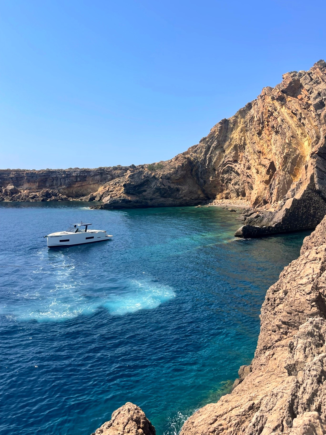 Guests anchored below the pine cliffs of Cala Llamp, the water turquoise and still.